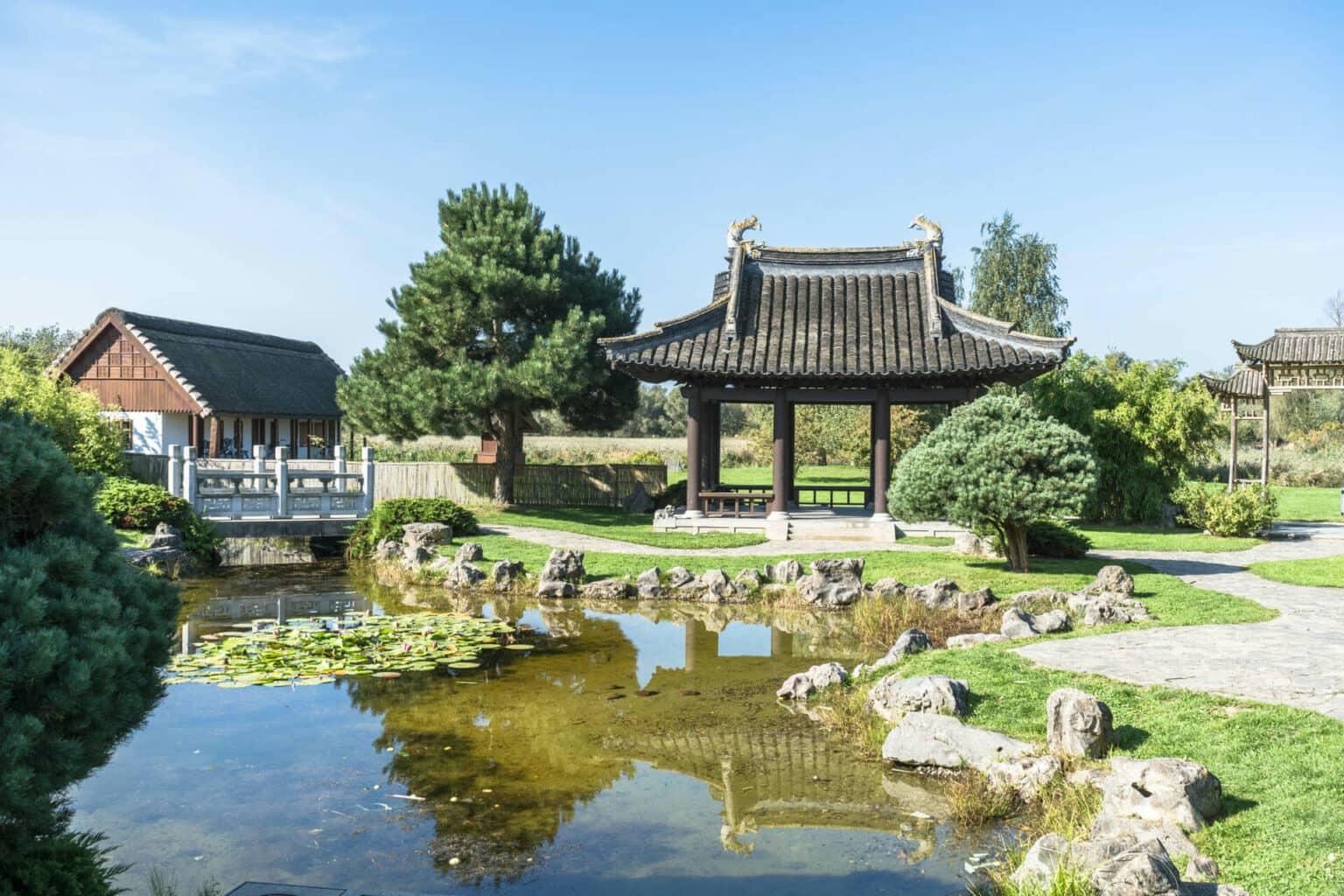 In the Japanese Garden in Rostock, two traditional temples stand behind a small pond adorned with sea leaves, over which a white traditional bridge leads.