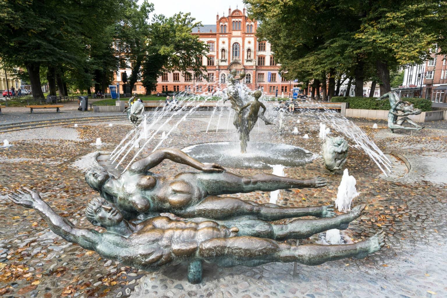 The Porno Fountain, consisting of four statues depicting couples in lovemaking, is located in front of an old red brick building, water sprays from the fountains.