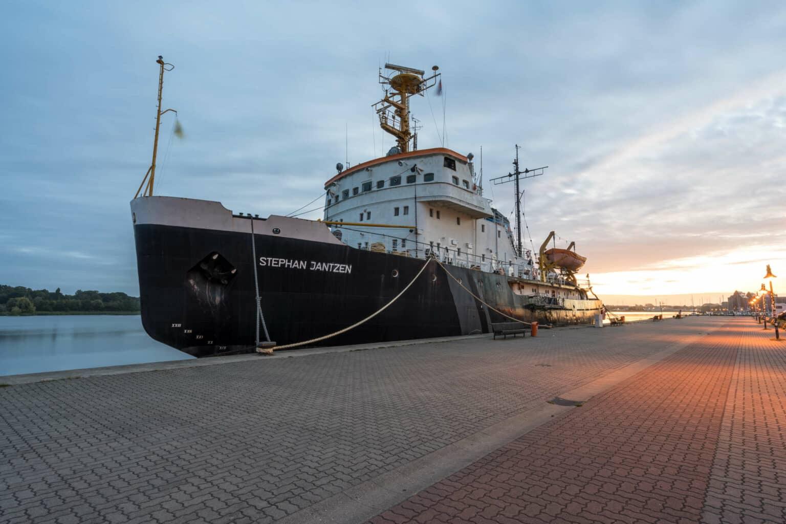 The Stephan Jantzen, a huge icebreaker, is docked in Rostock harbor, with the sun rising in the background, the ship is moored to a cobblestone platform lined with streetlights.
