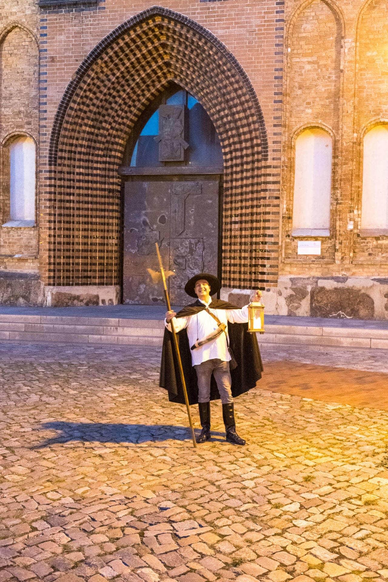 A night watchman stands in medieval costume with an oil lamp in hand in front of a large church door in Rostock. He shows visitors around the city.