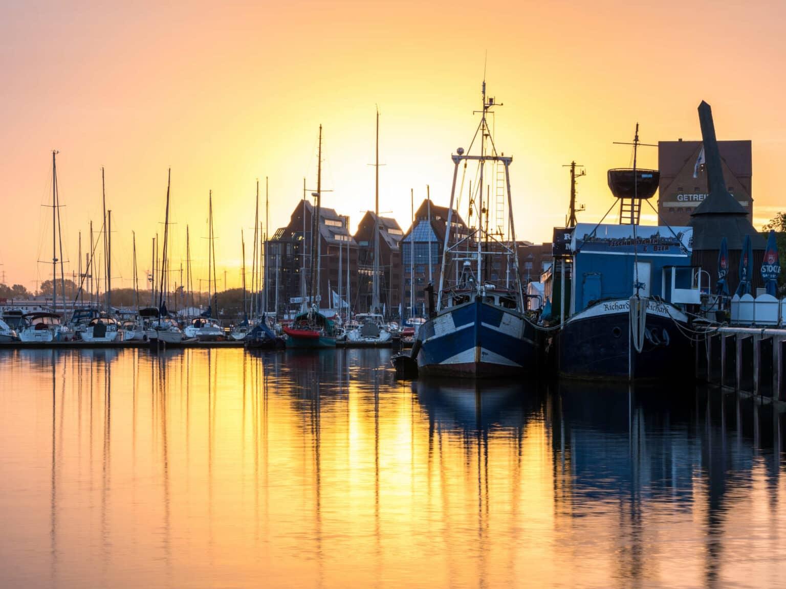Various ships, yachts, and steamers are docked at the Rostock city harbor at sunrise, the setting sun reflects in the water.