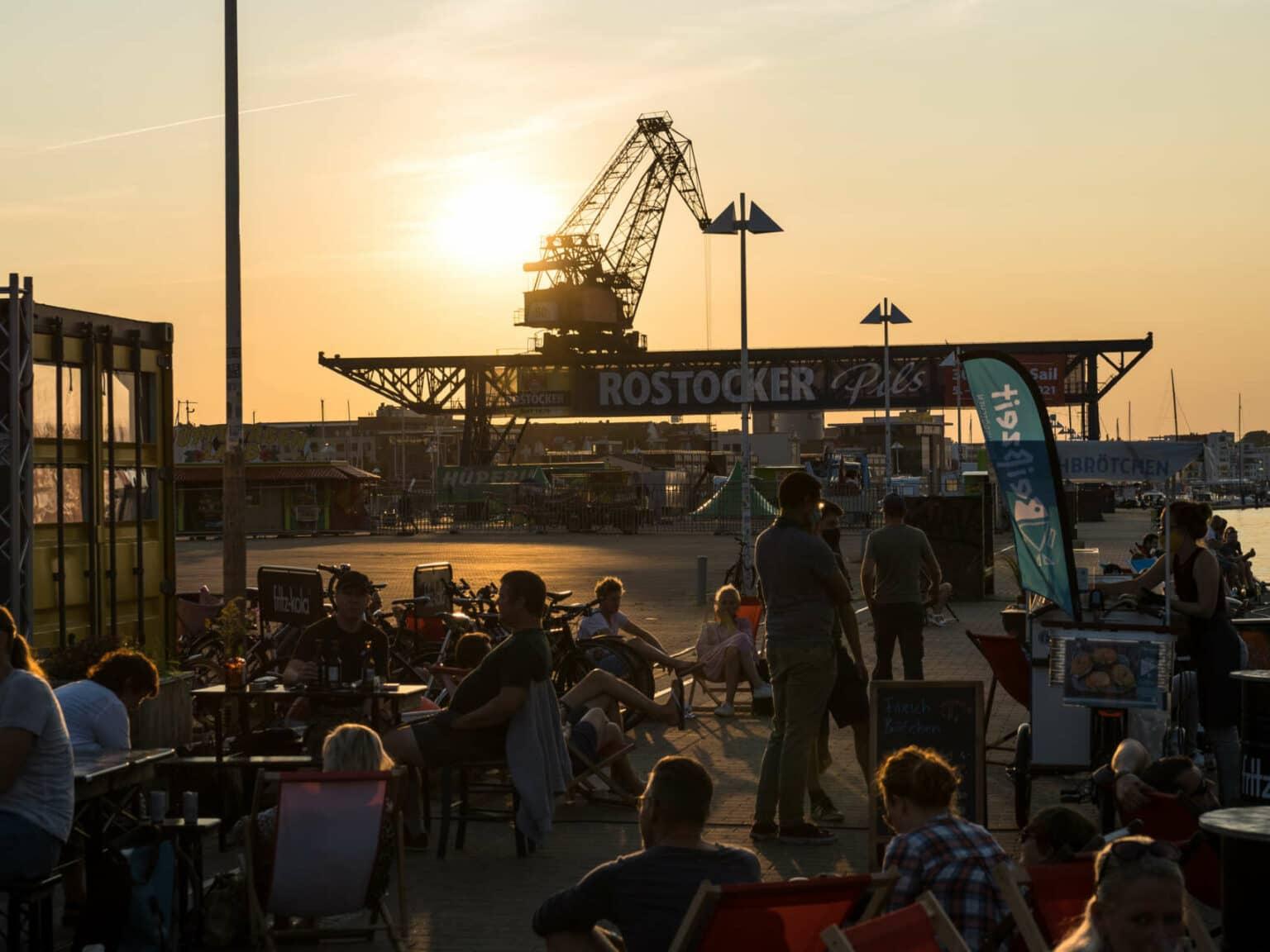 In summer, people are sitting in a harbor bar in Rostock at sunset, with a ship crane in the background.
