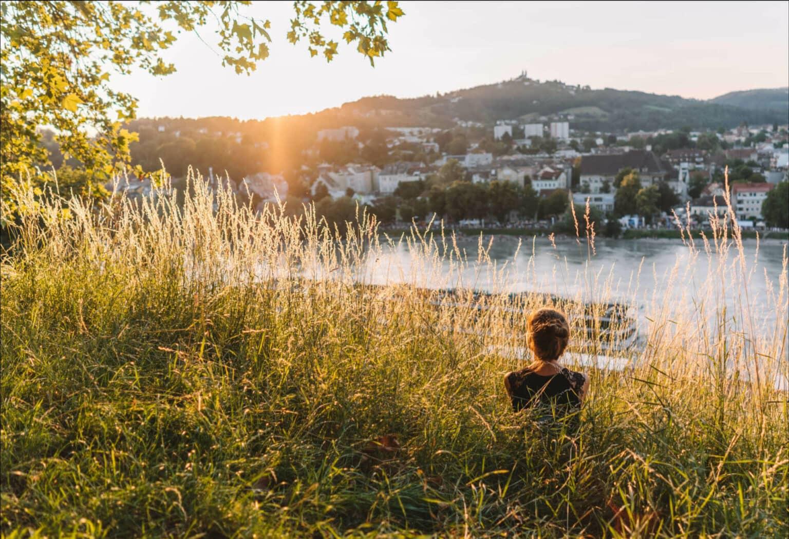 La bloguera de viajes Melanie se sienta en la hierba alta y disfruta del atardecer en el Danubio, que fluye por la impresionante ciudad de Linz.