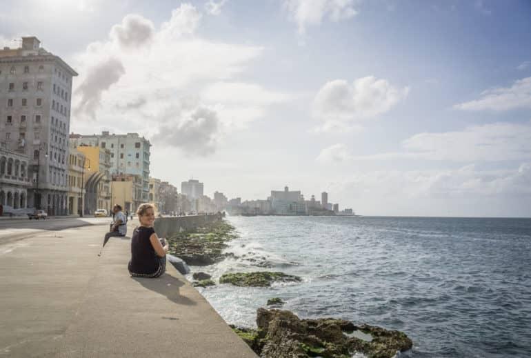 Una mujer se sienta en el Malecón al borde de la carretera que da al mar y sonríe a la cámara.