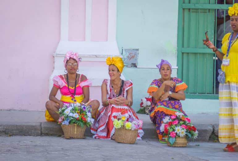 Tres mujeres en vistosos trajes cubanos se sientan con sus cestas de flores al borde de la carretera en La Habana; una mujer vestida de amarillo se les une y fuma en pipa.