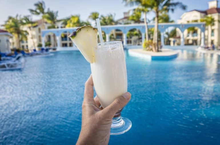 Una piña decora el vaso de cóctel lleno de Piña Colada que se sostiene frente a la cámara, al fondo se extiende el paisaje de piscina de un hotel.
