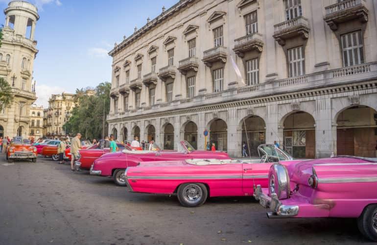 Varias coches antiguos de diferentes tonos de rojo se encuentran frente a un bloque de casas en La Habana, Cuba.