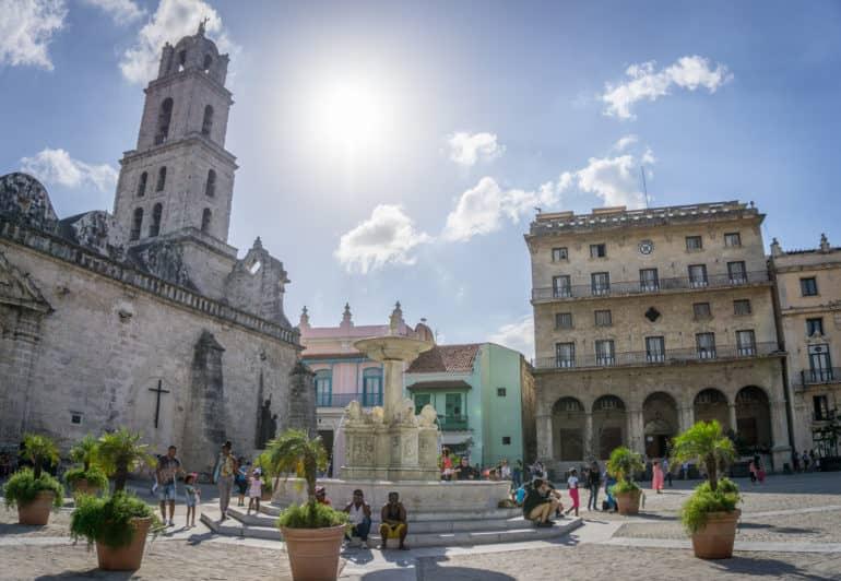 Vista de las fachadas de casas iluminadas por el sol; a la izquierda se ve una típica iglesia cubana y en medio de la plaza hay gente alrededor de una fuente.