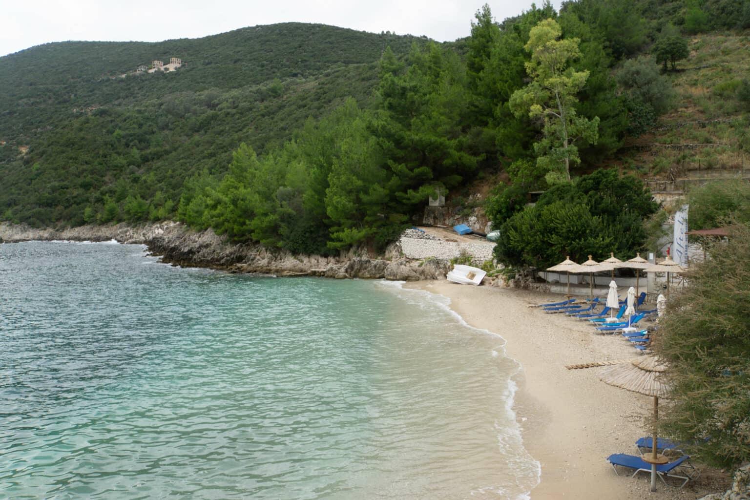 Blue sunbeds and straw umbrellas lie on the sandy beach of Afteli, the sea is turquoise with no waves, and a green, forested hillside rises over the bay.