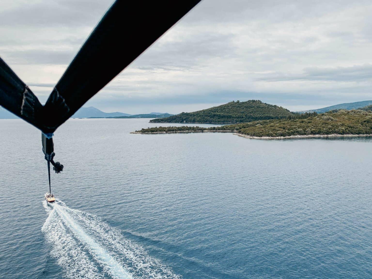 A parasailing boat leaves trails in the sea, above it the wings of the parasailing device unfold, with the extensive sea and the outskirts of Lefkada in the background.