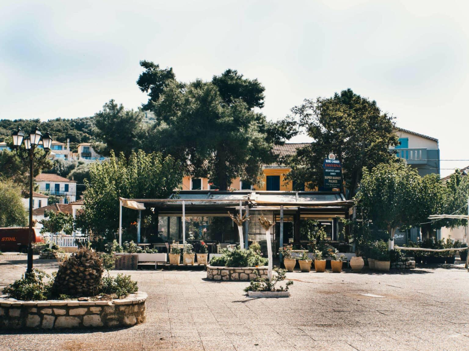 A cobbled square on the island of Meganisi, a yellow house is visible behind many green trees.