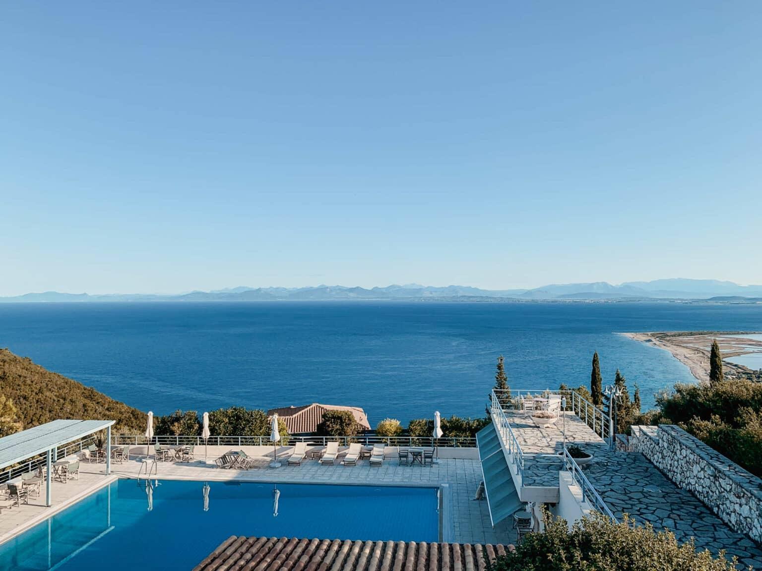 The pool at Mira Resort with sun loungers and umbrellas as well as covered terraces offers a stunning view of the bay and the ocean on Lefkada, with mountains in the distance.