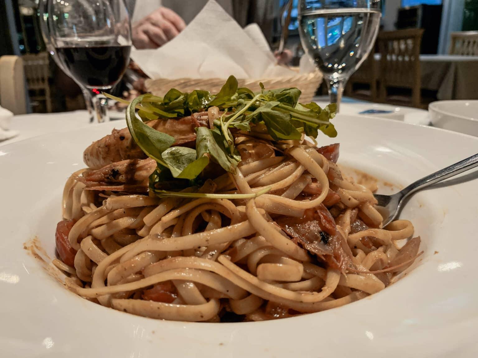 Culinary delights also impress visitors to Lefkada: On a plate, pasta with shrimp and arugula is presented, behind it are a water glass and a wine glass as well as a bread basket.