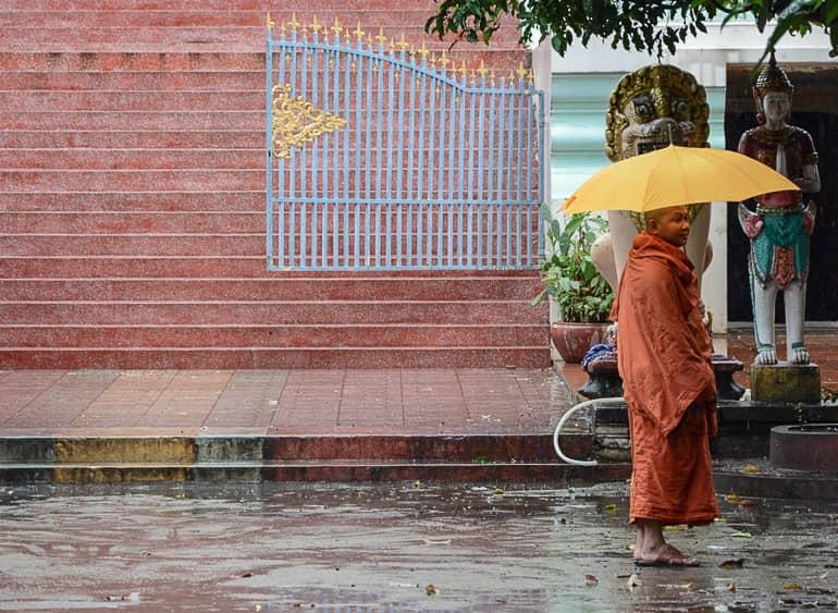 A monk in orange clothing and an umbrella walks barefoot on the rain-soaked streets in front of the walls of the Lotus Temple in Cambodia, Phnom Penh.