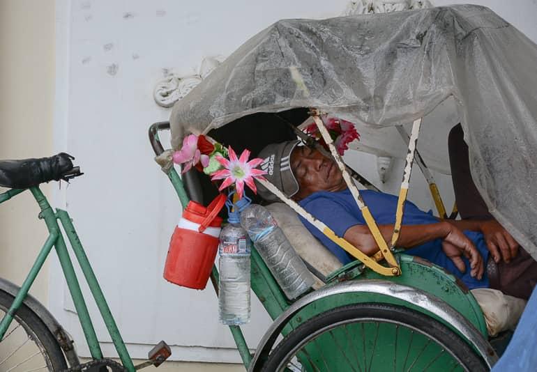 A cyclo driver rests against a house wall in the streets of Phnom Penh in his green rickshaw, with drinks and flowers attached to the handlebars.