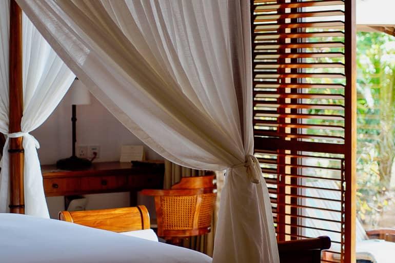 En una de las villas de The Residence Zanzibar en Tanzania, cortinas blancas decoran la cama con dosel, mientras que el escritorio y la silla al fondo son de madera de caoba y la vista a través de la puerta del patio muestra una tumbona de descanso.