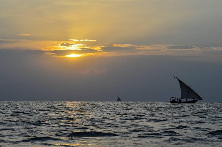En el mar de Tanzania, el sol se oculta detrás de las nubes mientras pequeños dhows con velas navegan pacíficamente.