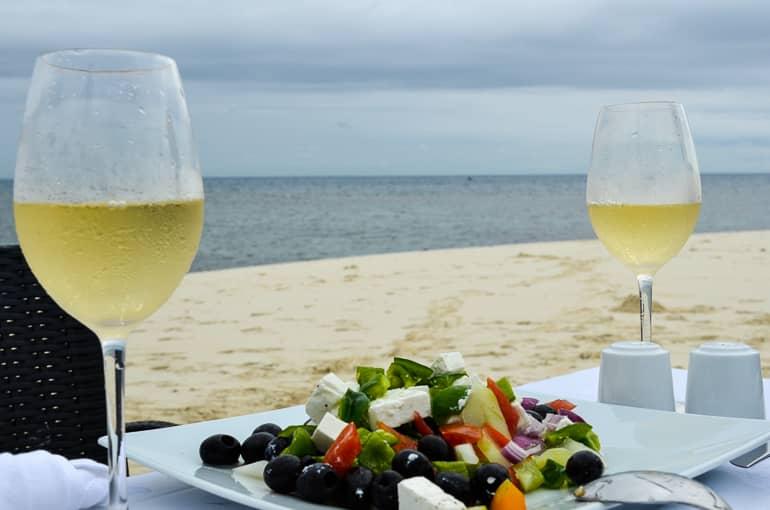 En una cena romántica en la playa de Zanzíbar hay 2 copas de vino blanco y una ensalada servidas en la mesa.
