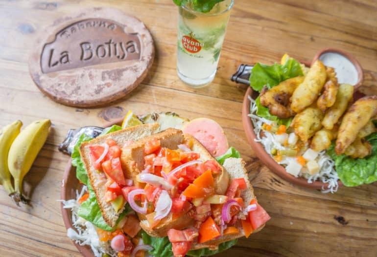 En la mesa de madera de la Taberna La Bojita en Trinidad, Cuba, hay dos platos con ensalada y tostadas, además de un mojito y plátanos baby.