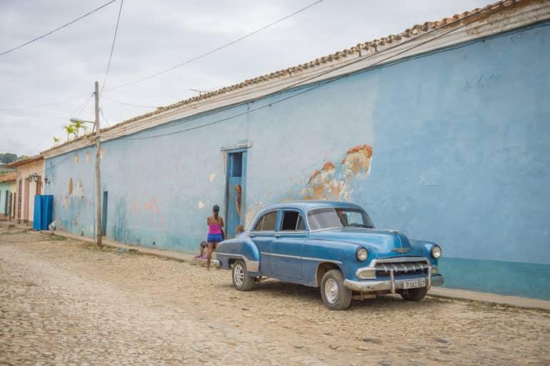 En una calle de Trinidad en Cuba, hay un automóvil clásico azul frente a una fachada de casa azul, de cuya puerta azul un hombre asoma y habla con una niña en la calle.