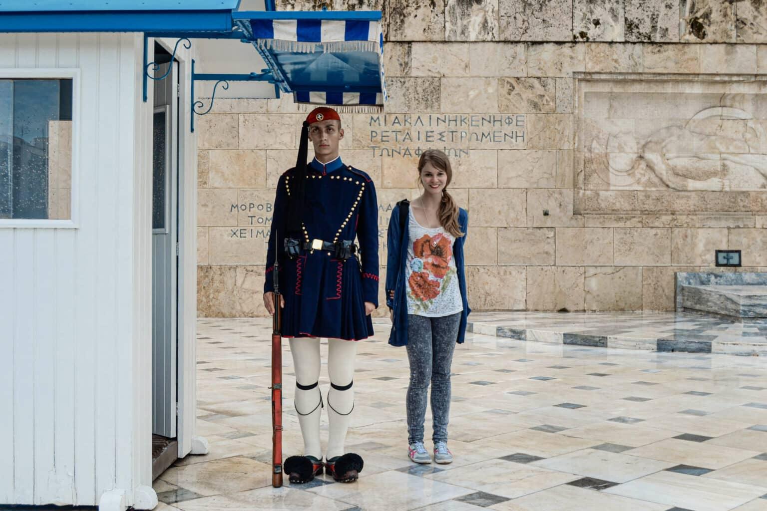 A guard in traditional costume in front of the Greek Parliament in Athens, beside him a smiling woman poses for the photo.