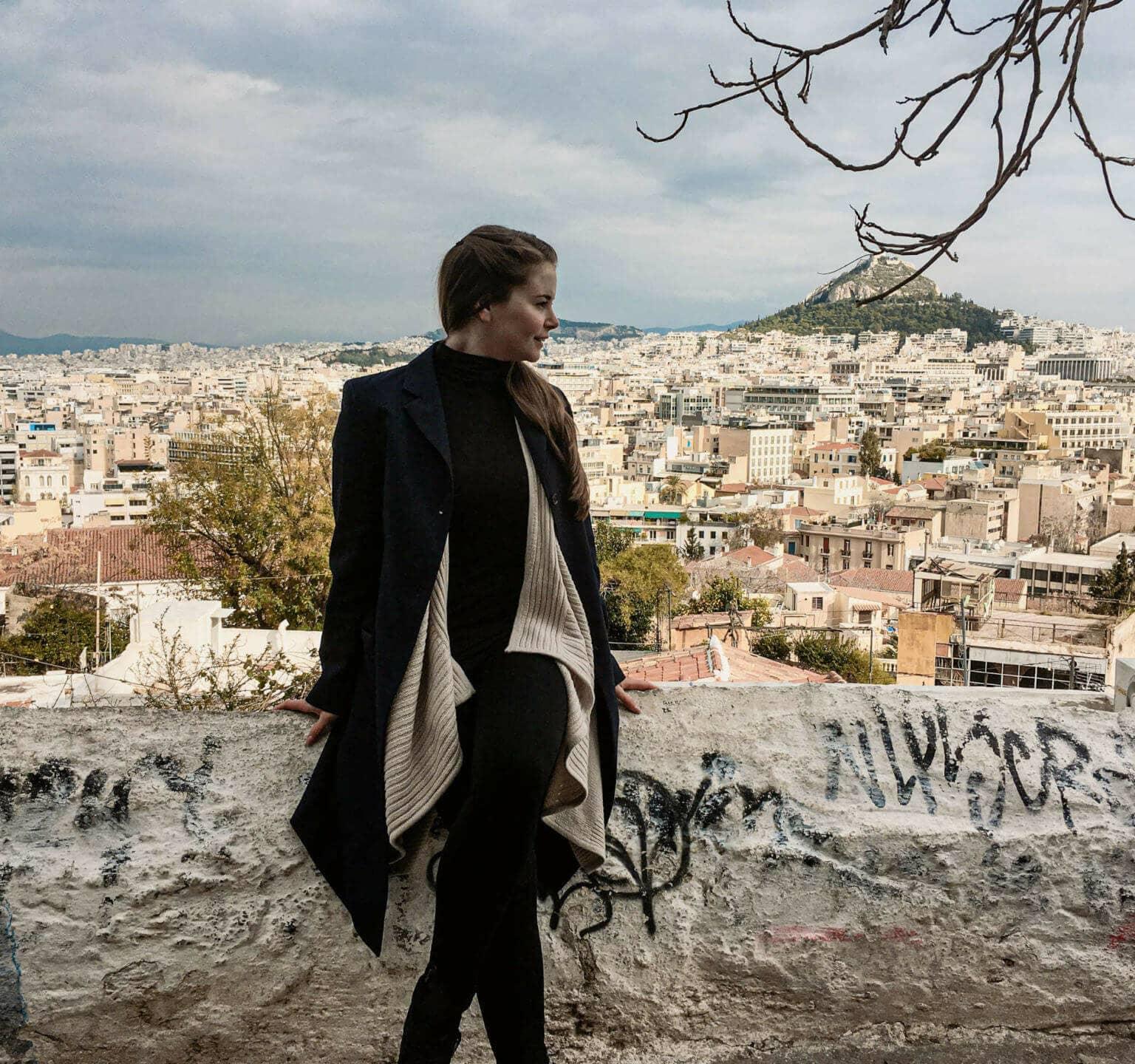 View of the sprawling capital city of Athens and the Acropolis from Anafiotika, travel blogger Franziska poses sitting on a wall overlooking the rooftops.
