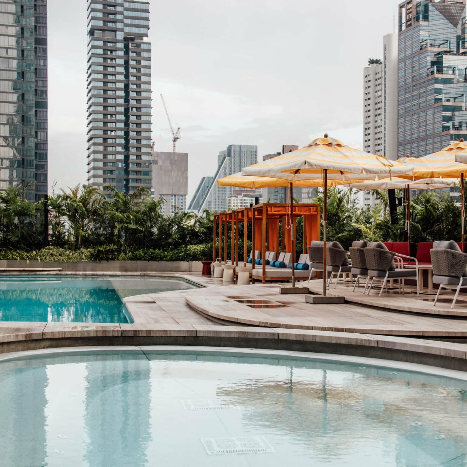 A large hotel pool in the foreground surrounded by palm trees and skyscrapers in Bangkok.