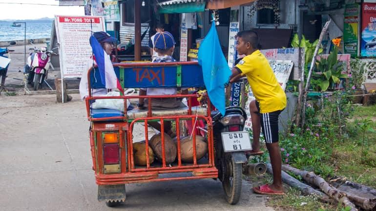 Niños locales conversando después de hacer compras junto a un taxi motorizado en Koh Mook, Tailandia.