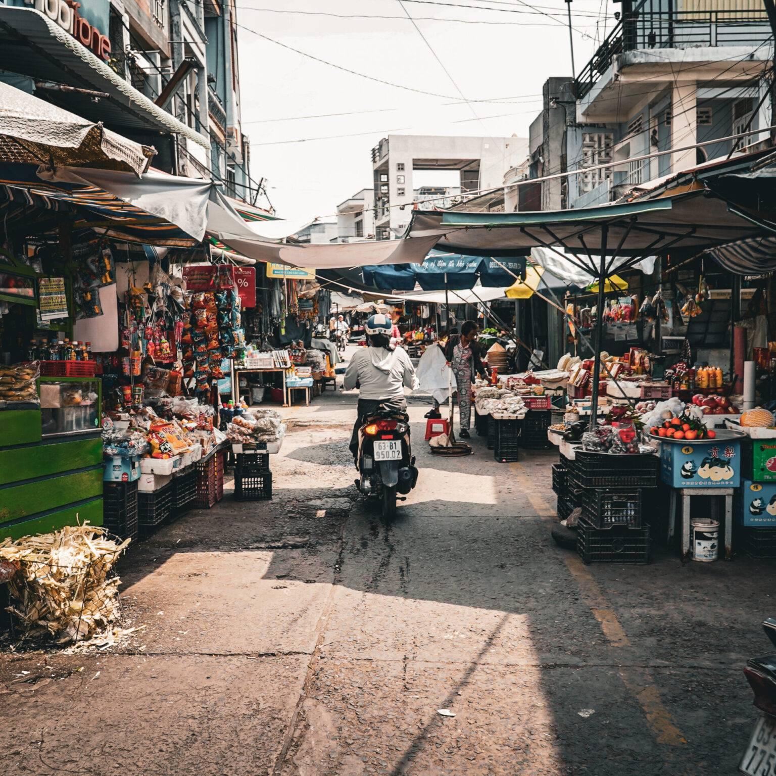 A scooter rides through the Cai Be market with colorful stalls.
