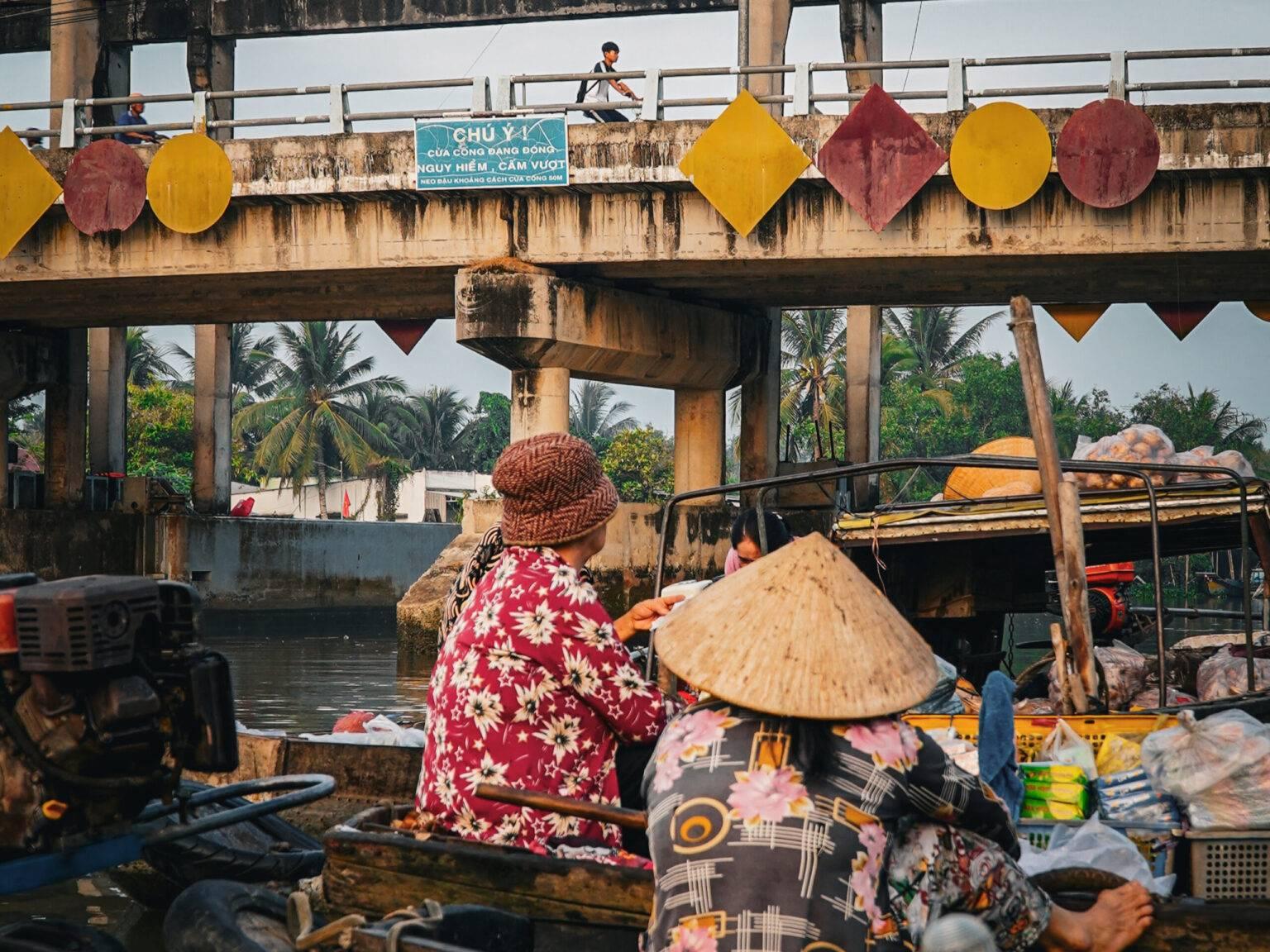 People at the Phong Dien floating market under a bridge on wooden boats.