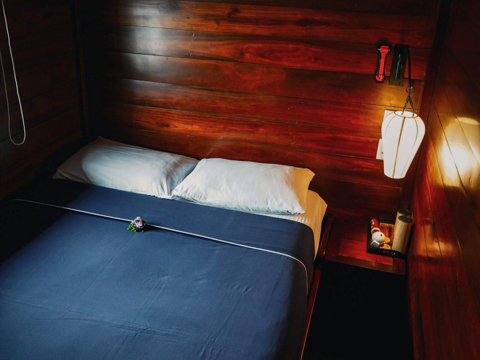 A cabin on the Basaac 2 with a double bed, a nightstand with a lamp, and walls paneled with dark wood.
