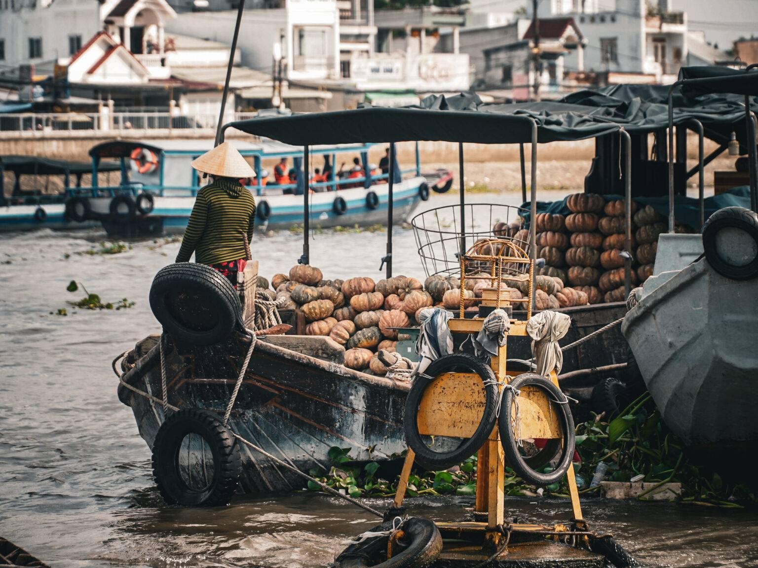 A man with a pointed straw hat sits on a boat loaded with pumpkins.