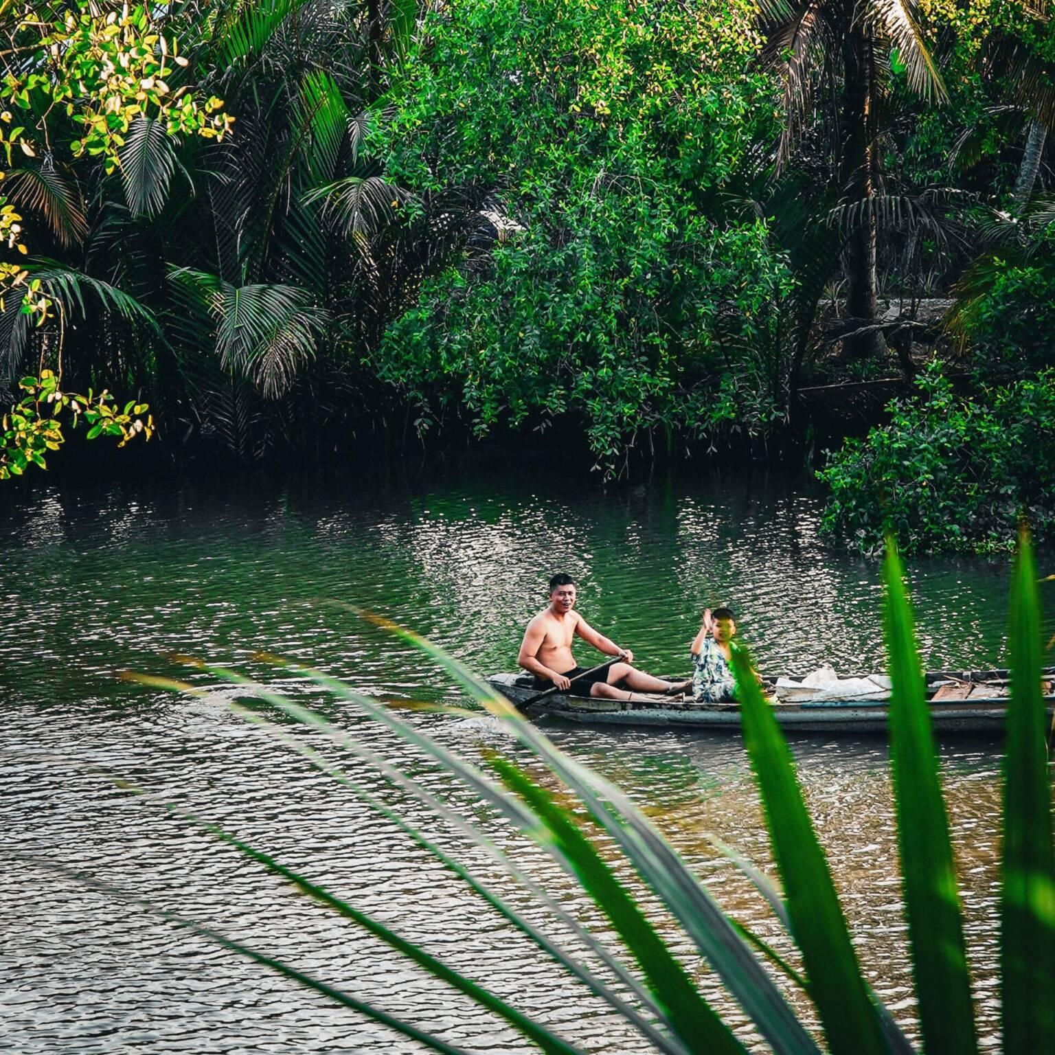 Father and son float on a long boat along a river surrounded by green plants.