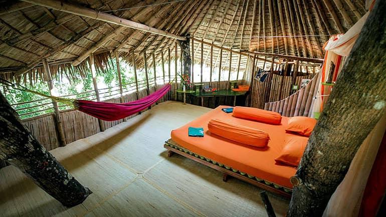 En el interior de la casa del árbol del Bukat Raya en Borneo hay una cama de madera con fundas naranjas y una hamaca morada.