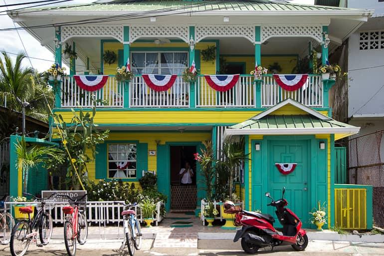 Delante de una casa amarilla-verde de estilo colonial, ricamente decorada con plantas y banderas, hay un scooter rojo en Bocas del Toro, Panamá.