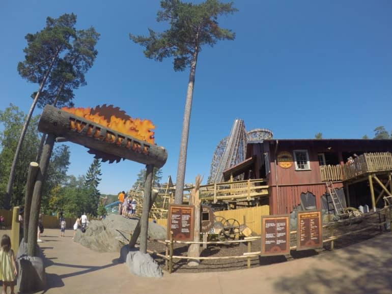 Al lado de la entrada del parque de atracciones Wildfire Rollercoaster en Suecia hay una cabaña de madera marrón, al fondo hay una montaña rusa.