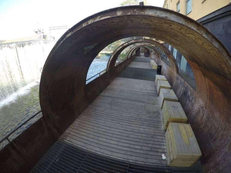 Un puente con encanto industrial, flanqueado por arcos de hierro oxidados y cajas de madera con vista al río en Norrköping, Suecia.