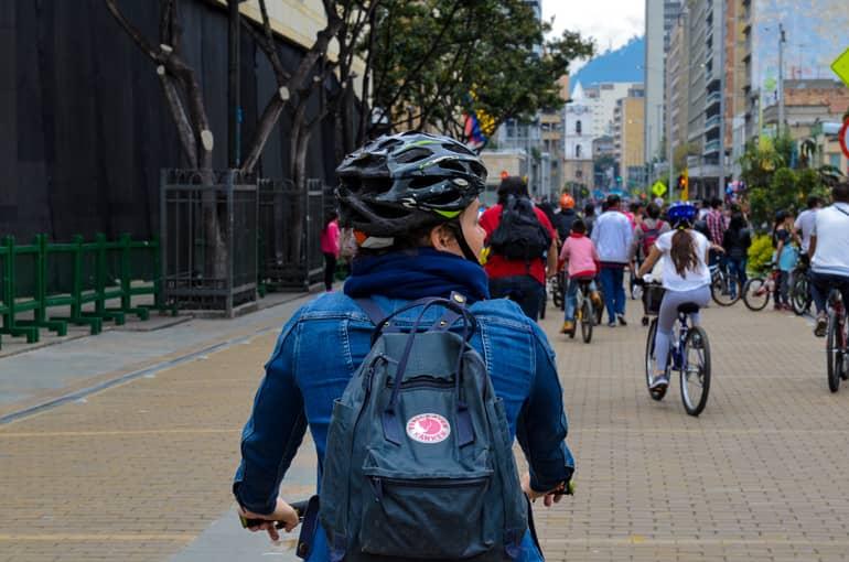 On a street in Bogotá, cyclists are out, with a female cyclist wearing a blue outfit and carrying a backpack glancing sideways.