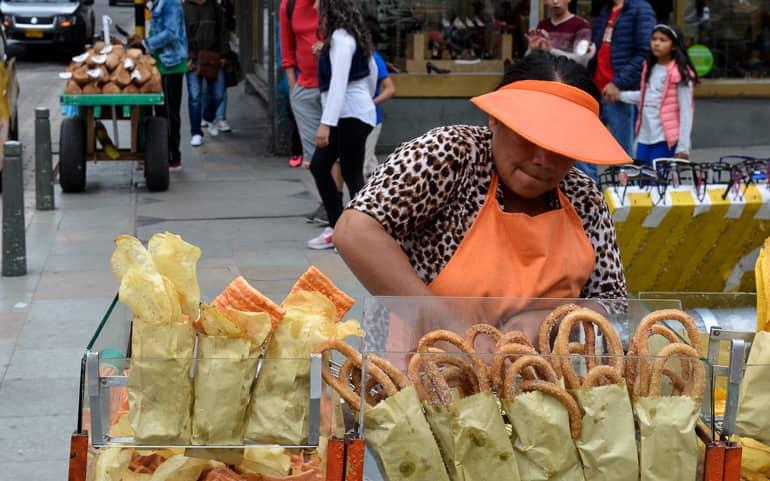 On the streets of Bogotá, a local woman in an orange apron and cap sells fried pastries.