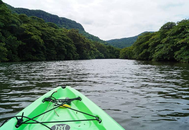 A green kayak floats on the river of Iriomote, one of the Yaeyama islands of Japan surrounded by dark green hills.