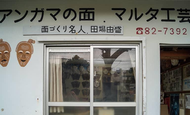 The small house facade with a double window and open door, as well as blue Japanese writing indicates the office of mask maker Mr. Taba on the Yaeyama island of Ishigaki in Japan.