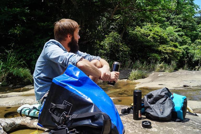 The blogger Clemens sits near the Sangara Waterfalls in Iriomote, Japan between backpacks on stones in the water and holds an open thermos in his hand.