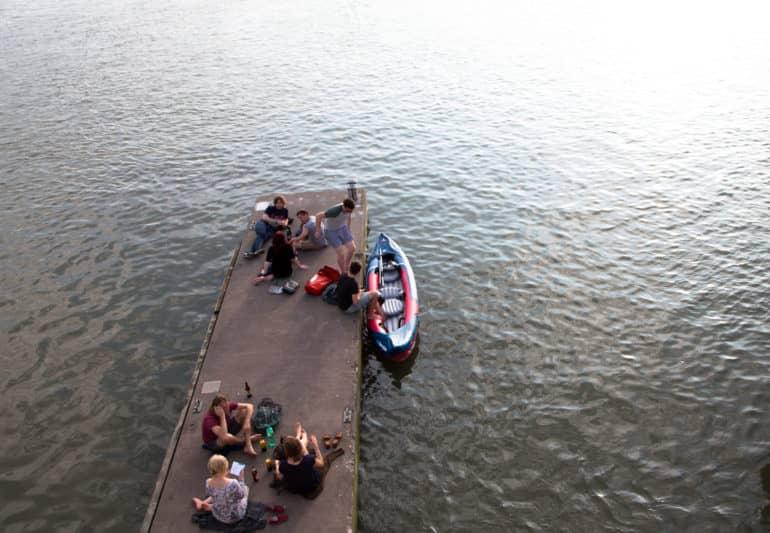 Young people sit on a dock by the Bristol harbor, chatting, while two individuals are getting into a kayak at the side of the dock.