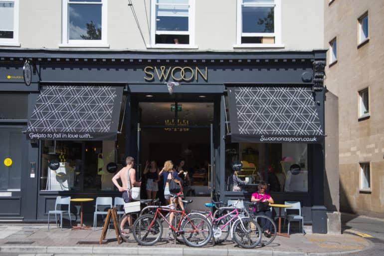 People walk towards the entrance of Café Swoon Gelato in Bristol, with its dark house facade, black awnings, and golden lettering.