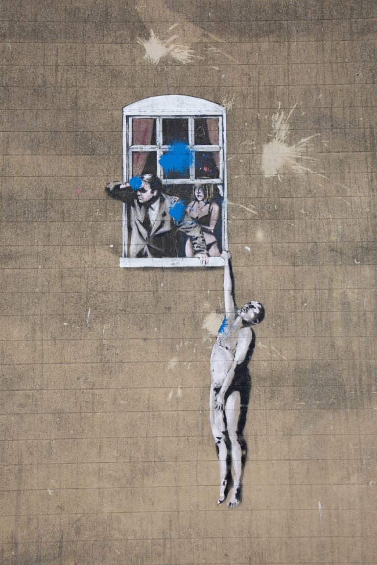 On a building facade in Bristol, there's an artwork in Banksy's style - a couple is drawn, with a man looking out the window with binoculars, while the woman anxiously reaches for his arm in her underwear, as another man holds on to the windowsill nude with one arm.