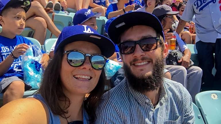 Una pareja sonriente mira a la cámara durante un partido de béisbol de los L.A. Dodgers, ambos llevan gorras azules de L.A.