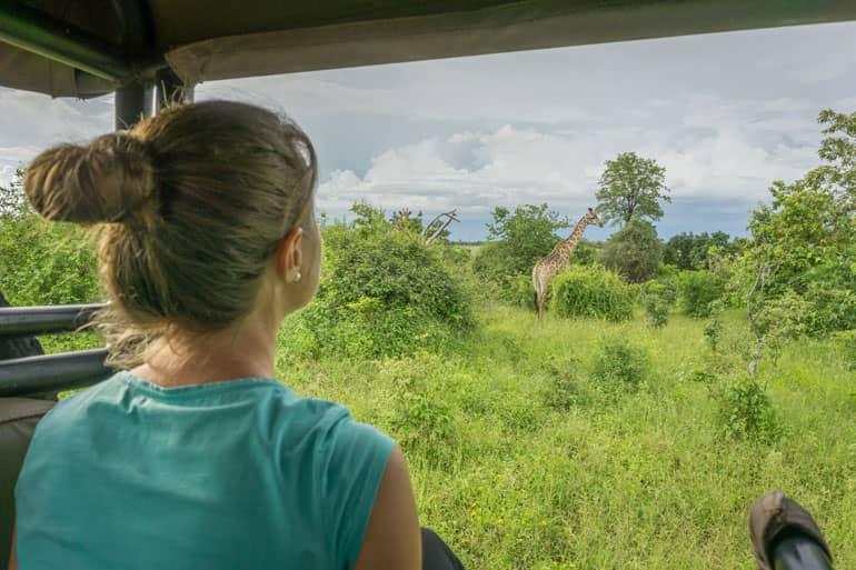 A woman faces another woman turned away from the camera in Chobe National Park, with a giraffe in a meadow in the background.