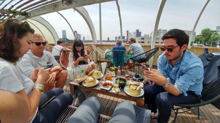 En la terraza del Aloha Bar, jóvenes están sentados alrededor de una pequeña mesa llena de comida, algunos miran concentrados sus teléfonos.