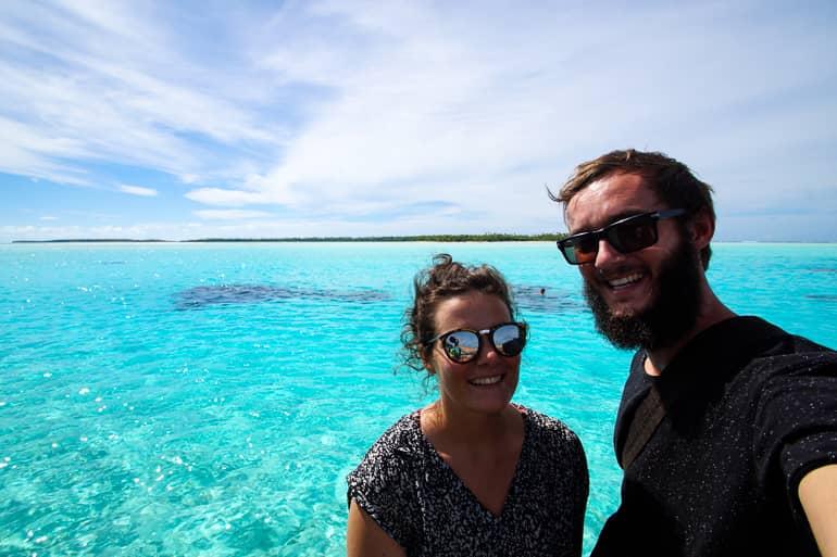 The two bloggers Anne and Clemens smile brightly at the camera from a Vaka on the light blue sea of the Cook Island Aitutaki.